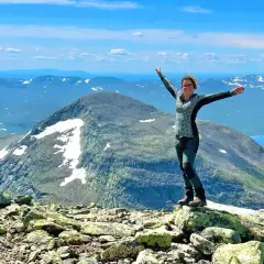 Kvinna med armarna utsträckta på en stenig topp, njuter av utsikten över Sveriges berg och sjöar under en klarblå himmel.