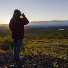 Kvinna i rutig skjorta fotograferar vid midnattssol i Sverige. Vackert landskap med sjöar och skog i gyllene ljus.