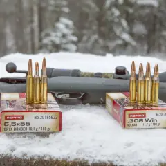 Två lådor med Norma ammunition, 6,5x55 och .308 Win, står på snö framför ett gevär. Klass 1 ammunition redo för jakt.
