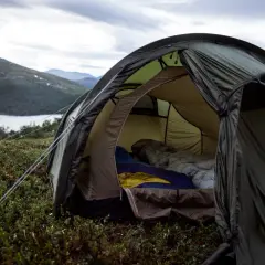 En öppen tält med sovsäckar redo för natten. En mysig känsla av värme i tält utan el, med en sjö och berg i bakgrunden.