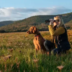Kvinna med kikare och en brun hund i ett fält. Perfekt för kortdrivande hundraser som Vizsla.