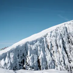En grupp vandrare på en snötäckt bergstopp under en helags topptur. Den isiga bergssidan kontrasterar mot den klara blå himlen.