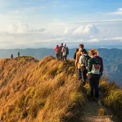 A group of hikers with backpacks walk along a grassy mountain ridge with a scenic view of rolling hills.