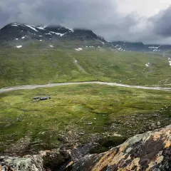 Grön dal med en flod och fjällstugor, som en karta över Kungsleden. Moln hänger över snöklädda berg.