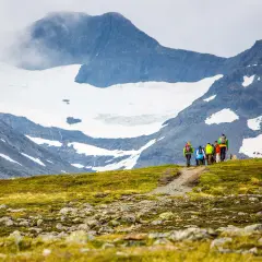 Vandringsgrupp med ryggsäckar på en stig i fjällen, med snöklädda toppar och moln som omger sylarna runt.
