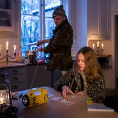 Två personer i ett kök med levande ljus och en sprit kamin. En flicka spelar kort vid ett bord, medan en person lagar mat vid spisen.