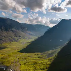 En dramatisk dalgång längs Kungsleden, med solljus som bryter igenom molnen och kastar långa skuggor över den gröna, mossbeklädda marken.