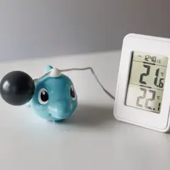 En blå delfin med en svart kula och en digital termometer som visar 21.6°C utomhus och 22.1°C inomhus. Undrar hur kallt kan man ha i ett hus?