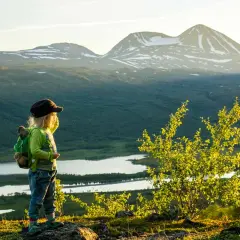 Ett barn med ryggsäck blickar ut över en vidsträckt fjällvy med sjöar och snöklädda toppar. En perfekt bild av vandring med boende i naturen.