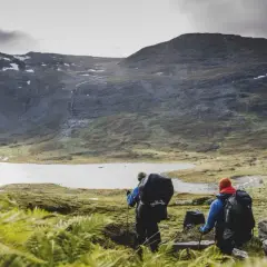 Två vandrare med ryggsäckar går mot en sjö i fjällen. En karta över midnattssolens utbredning kan hjälpa dem att planera sin tur.