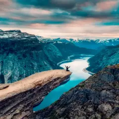 En person sitter på Trolltungan i Norge, med armarna utsträckta mot den dramatiska fjällnaturen och en glittrande sjö nedanför.