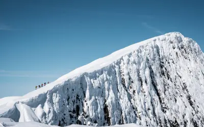 En grupp vandrare på en snötäckt bergstopp under en helags topptur. Den isiga bergssidan kontrasterar mot den klara blå himlen.