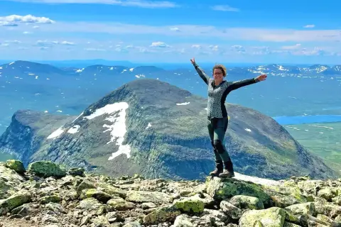 Kvinna med armarna utsträckta på en stenig topp, njuter av utsikten över Sveriges berg och sjöar under en klarblå himmel.