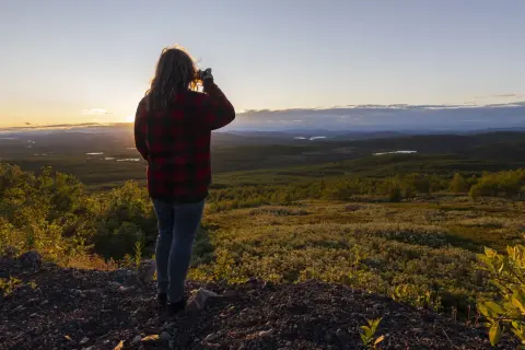Kvinna i rutig skjorta fotograferar vid midnattssol i Sverige. Vackert landskap med sjöar och skog i gyllene ljus.