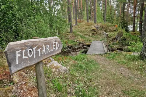 Träskylt visar vägen till flottarleden i Munkedal. En bro leder över en bäck i en skogsmiljö.