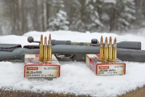 Två lådor med Norma ammunition, 6,5x55 och .308 Win, står på snö framför ett gevär. Klass 1 ammunition redo för jakt.