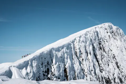 En grupp vandrare på en snötäckt bergstopp under en helags topptur. Den isiga bergssidan kontrasterar mot den klara blå himlen.