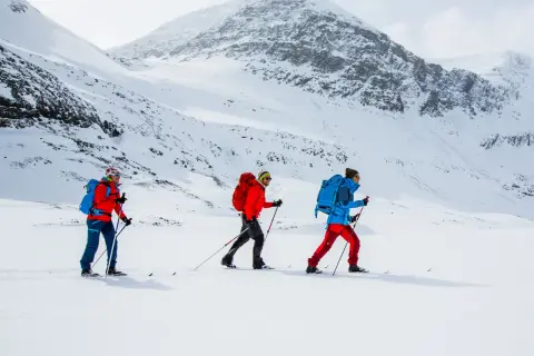 Tre personer åker skidor i ett snöigt landskap nära Abisko. De är klädda i färgglada jackor och ryggsäckar, redo för äventyr.