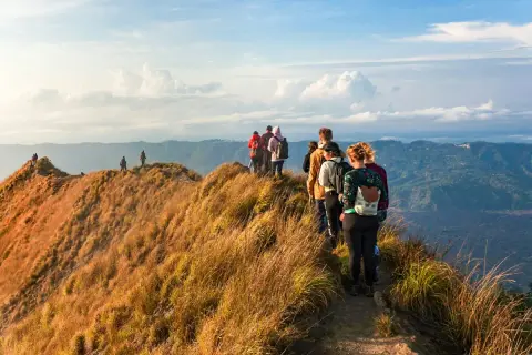 A group of hikers with backpacks walk along a grassy mountain ridge with a scenic view of rolling hills.