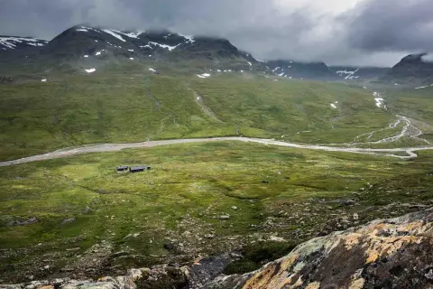 Grön dal med en flod och fjällstugor, som en karta över Kungsleden. Moln hänger över snöklädda berg.