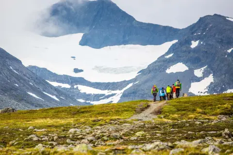 Vandringsgrupp med ryggsäckar på en stig i fjällen, med snöklädda toppar och moln som omger sylarna runt.
