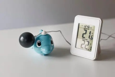 En blå delfin med en svart kula och en digital termometer som visar 21.6°C utomhus och 22.1°C inomhus. Undrar hur kallt kan man ha i ett hus?