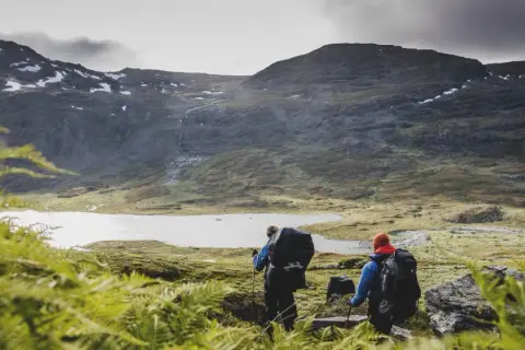 Två vandrare med ryggsäckar går mot en sjö i fjällen. En karta över midnattssolens utbredning kan hjälpa dem att planera sin tur.