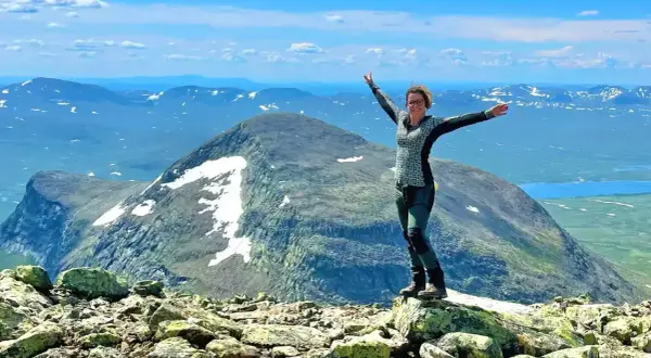 Kvinna med armarna utsträckta på en stenig topp, njuter av utsikten över Sveriges berg och sjöar under en klarblå himmel.