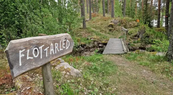 Träskylt visar vägen till flottarleden i Munkedal. En bro leder över en bäck i en skogsmiljö.