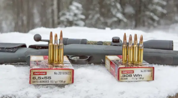 Två lådor med Norma ammunition, 6,5x55 och .308 Win, står på snö framför ett gevär. Klass 1 ammunition redo för jakt.