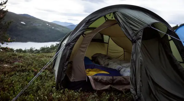 En öppen tält med sovsäckar redo för natten. En mysig känsla av värme i tält utan el, med en sjö och berg i bakgrunden.