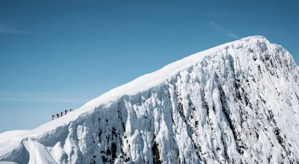 En grupp vandrare på en snötäckt bergstopp under en helags topptur. Den isiga bergssidan kontrasterar mot den klara blå himlen.