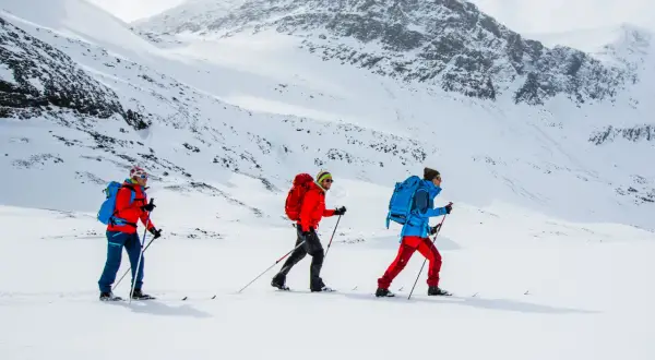 Tre personer åker skidor i ett snöigt landskap nära Abisko. De är klädda i färgglada jackor och ryggsäckar, redo för äventyr.