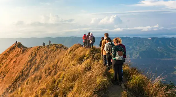 A group of hikers with backpacks walk along a grassy mountain ridge with a scenic view of rolling hills.