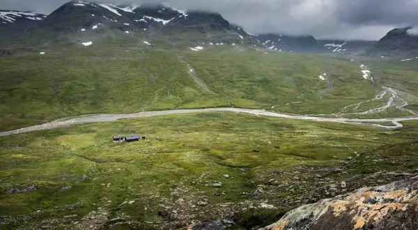 Grön dal med en flod och fjällstugor, som en karta över Kungsleden. Moln hänger över snöklädda berg.