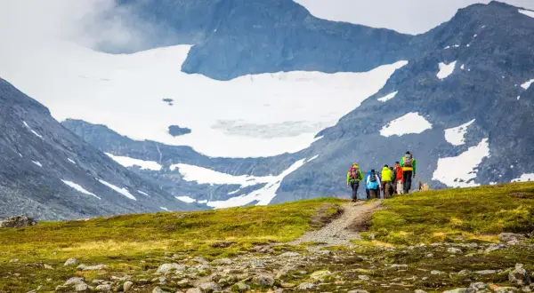 Vandringsgrupp med ryggsäckar på en stig i fjällen, med snöklädda toppar och moln som omger sylarna runt.