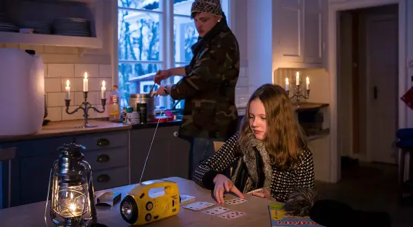 Två personer i ett kök med levande ljus och en sprit kamin. En flicka spelar kort vid ett bord, medan en person lagar mat vid spisen.