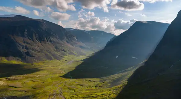 En dramatisk dalgång längs Kungsleden, med solljus som bryter igenom molnen och kastar långa skuggor över den gröna, mossbeklädda marken.