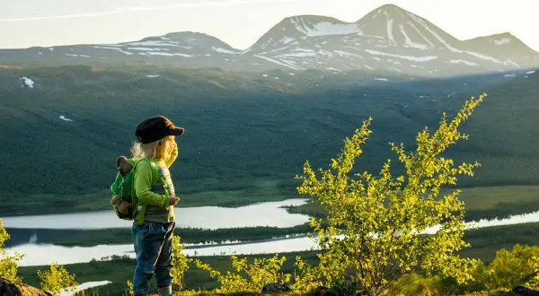 Ett barn med ryggsäck blickar ut över en vidsträckt fjällvy med sjöar och snöklädda toppar. En perfekt bild av vandring med boende i naturen.