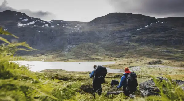 Två vandrare med ryggsäckar går mot en sjö i fjällen. En karta över midnattssolens utbredning kan hjälpa dem att planera sin tur.
