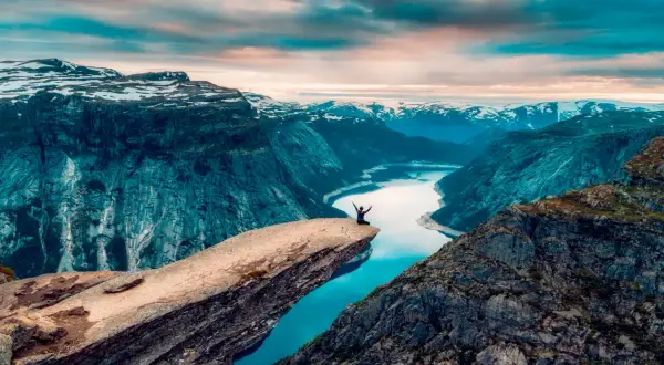 En person sitter på Trolltungan i Norge, med armarna utsträckta mot den dramatiska fjällnaturen och en glittrande sjö nedanför.