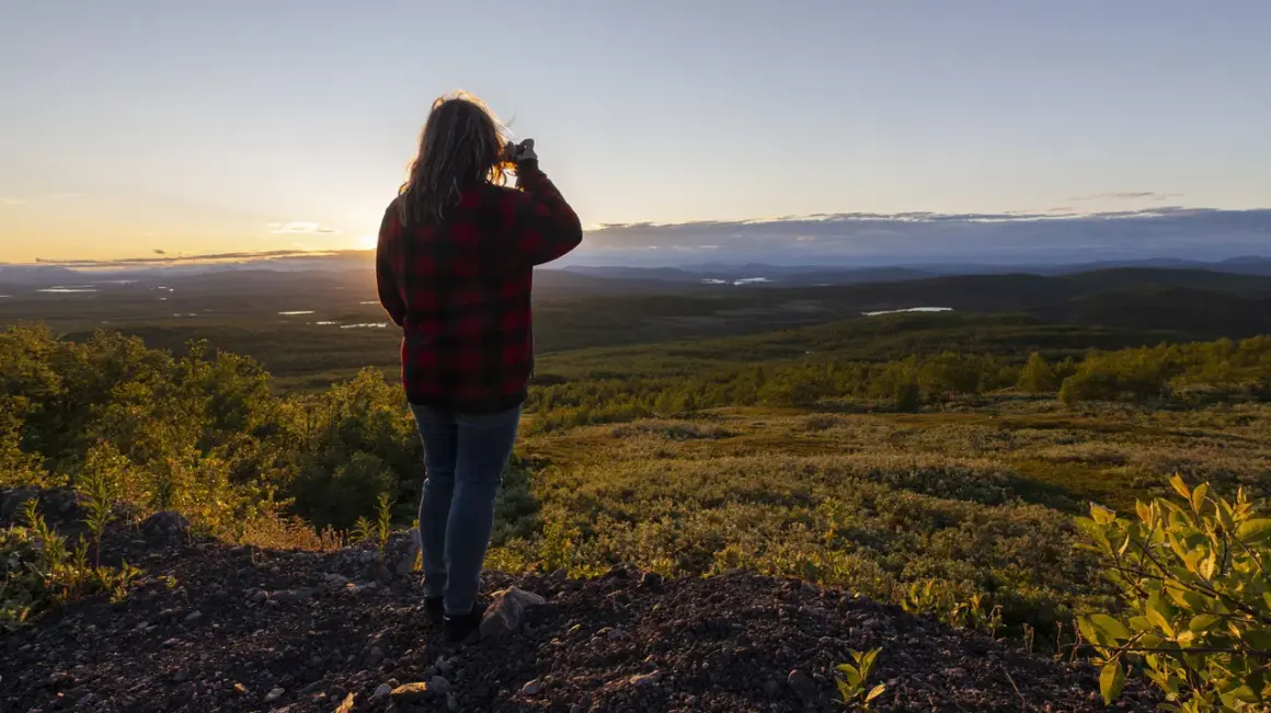 Kvinna i rutig skjorta fotograferar vid midnattssol i Sverige. Vackert landskap med sjöar och skog i gyllene ljus.