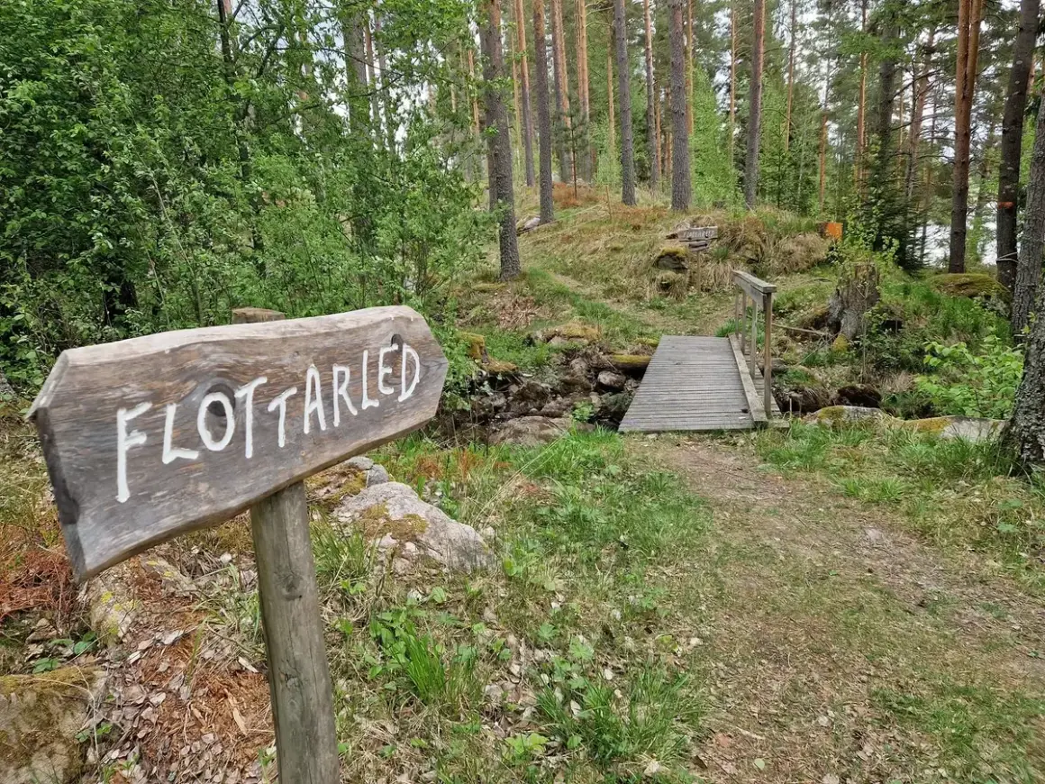 Träskylt visar vägen till flottarleden i Munkedal. En bro leder över en bäck i en skogsmiljö.