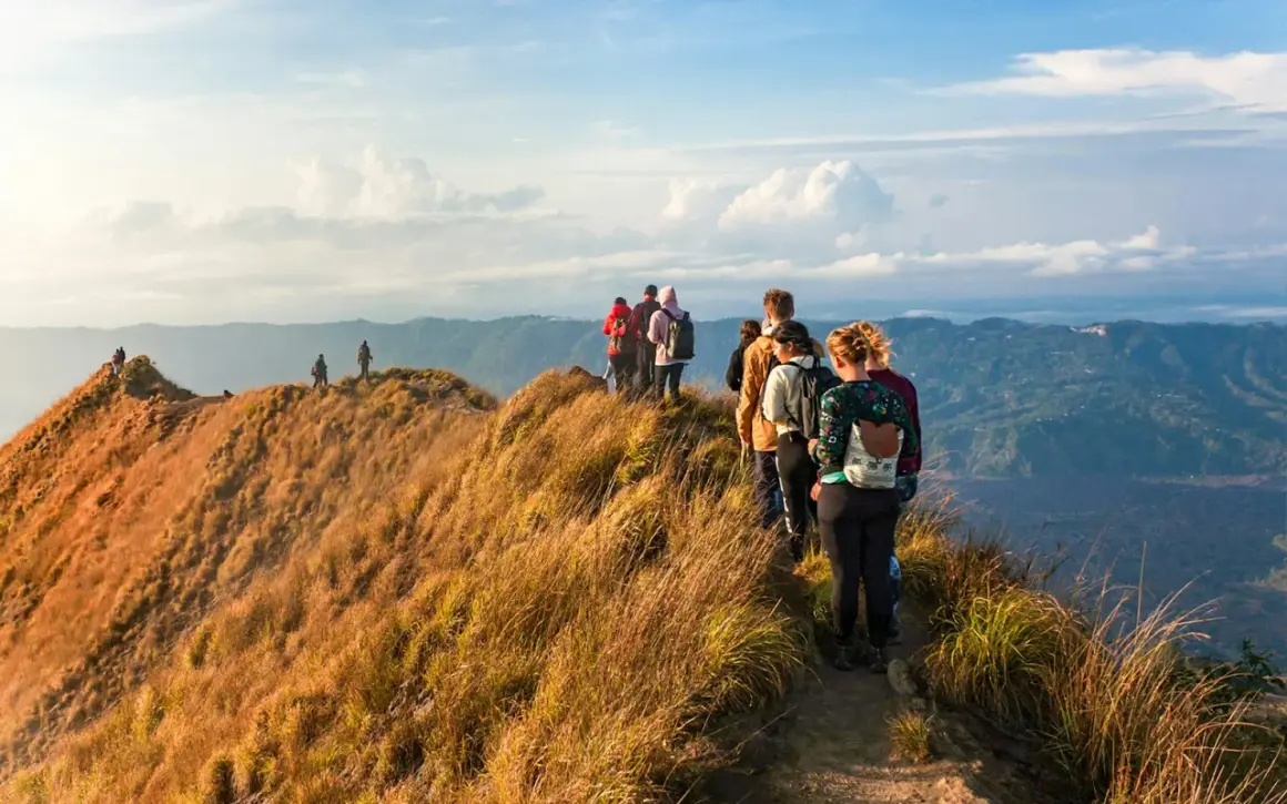 A group of hikers with backpacks walk along a grassy mountain ridge with a scenic view of rolling hills.