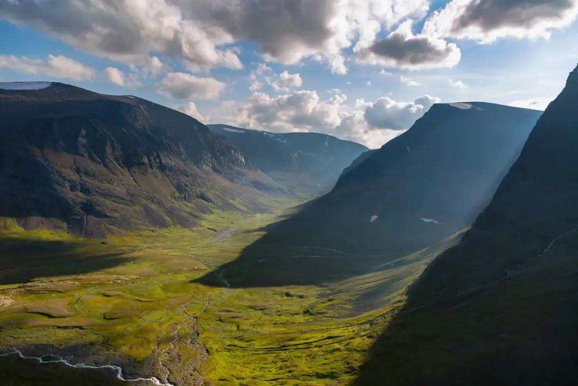 En dramatisk dalgång längs Kungsleden, med solljus som bryter igenom molnen och kastar långa skuggor över den gröna, mossbeklädda marken.