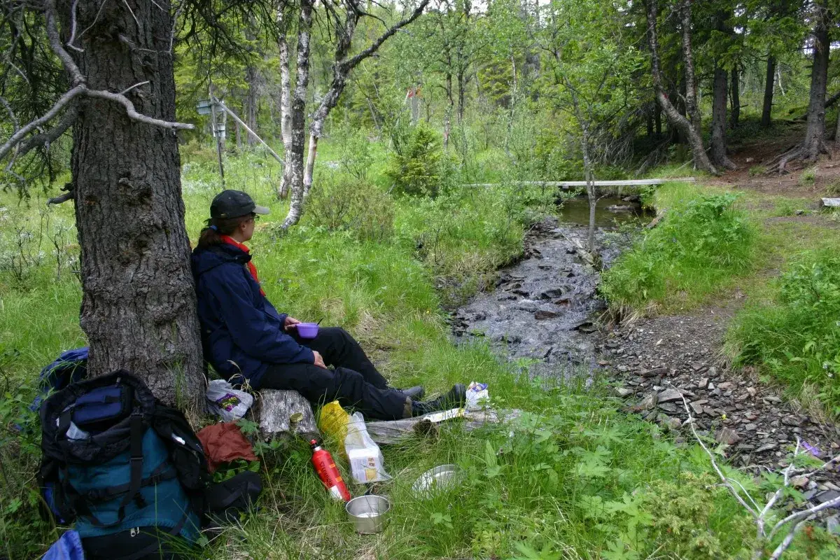 Vandrare vilar vid en bäck, med en ryggsäck och mat vid sidan. En liten bro syns i bakgrunden. Kanske har de precis passerat blå bandet.