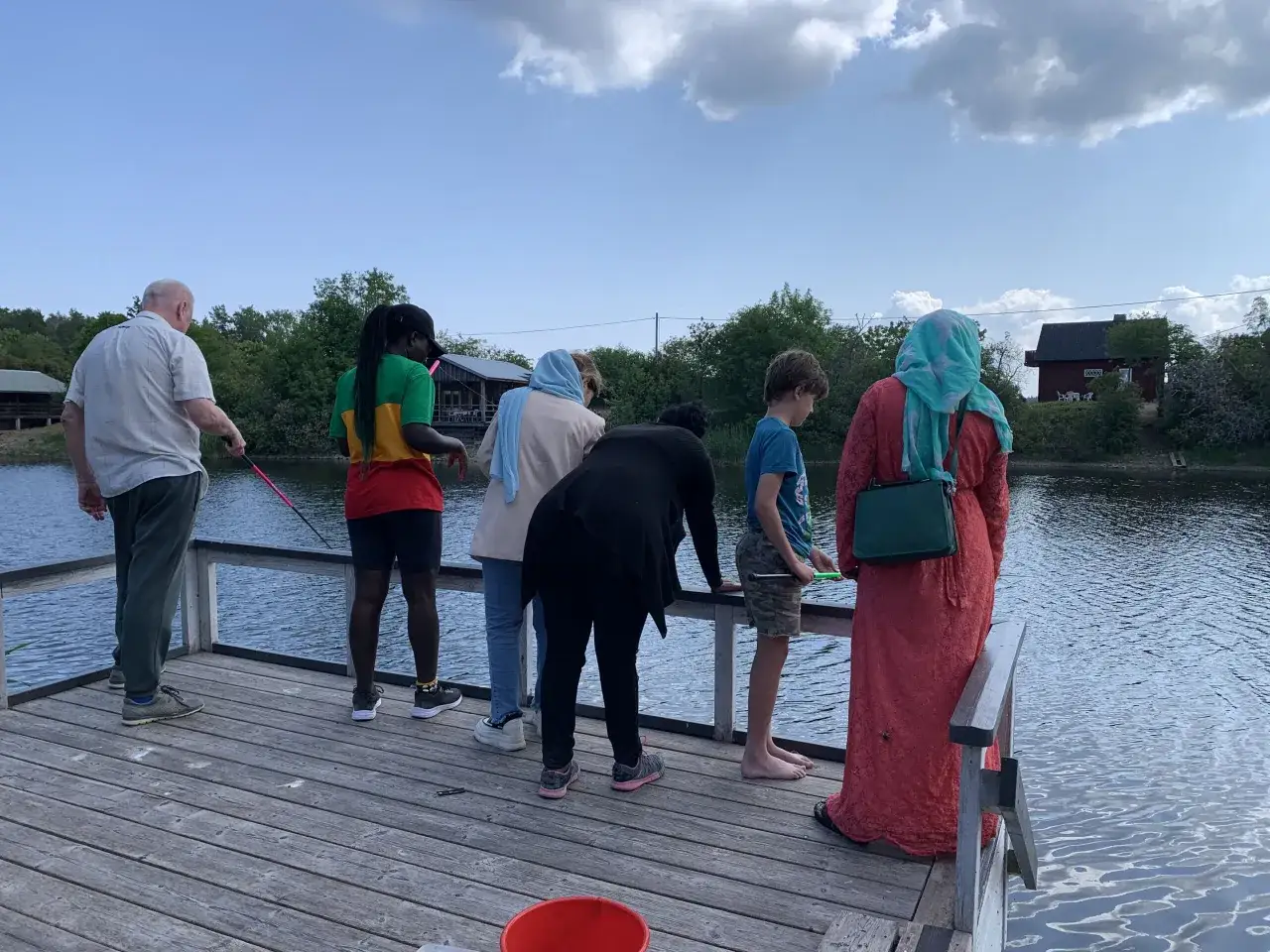 En grupp människor, inklusive en utter unge, fiskar från en brygga vid en sjö.