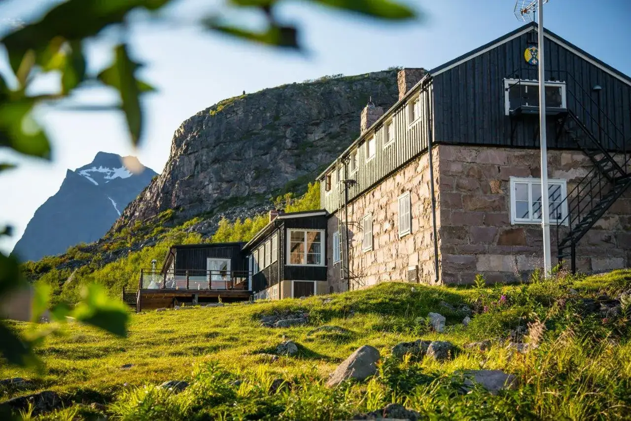 Boende vid Kebnekaise fjällstation, med stenbyggnad och mörka trädetaljer, omgivet av grönskande fjäll och en snöklädd topp i bakgrunden.