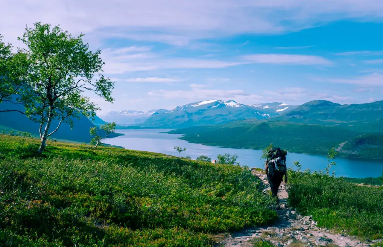 Vandrare med stor ryggs&auml;ck p&aring; en stenbelagd vandringsled norrland. Vacker sj&ouml; och sn&ouml;kl&auml;dda berg i bakgrunden.
