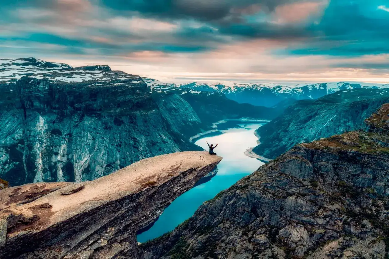 En person st&aring;r p&aring; Trolltunga, en ber&ouml;md klippavsats i Norge, med armarna utstr&auml;ckta mot den dramatiska fj&auml;llnaturen och en glittrande sj&ouml; nedanf&ouml;r.